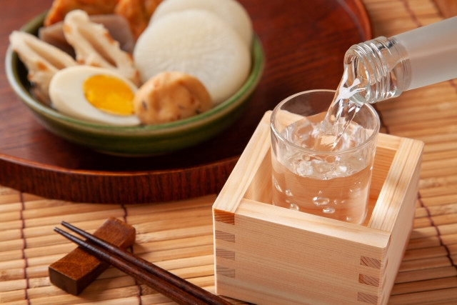Pouring of sake into glass placed inside masu with oden side dish